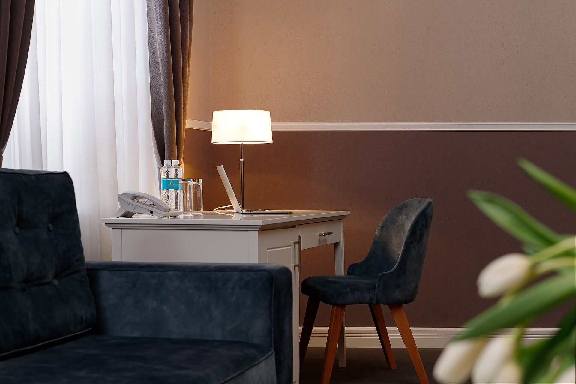 Une chambre d’hôtel avec un canapé bleu foncé, un bureau avec une lampe, une chaise, un téléphone et deux bouteilles d’eau.