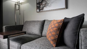Una sala de estar moderna con un sofá gris, una mesita de madera, un cojín naranja estampado, una lámpara de pie y un cuadro abstracto geométrico en la pared.
