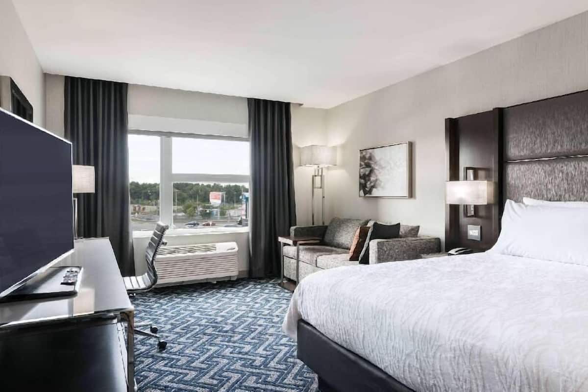 A hotel room with a bed, a desk with a TV, a sofa, a lamp, and a window with curtains.