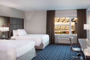 A hotel room with two beds, a desk, a chair, and a large window with a view of a building and trees.
