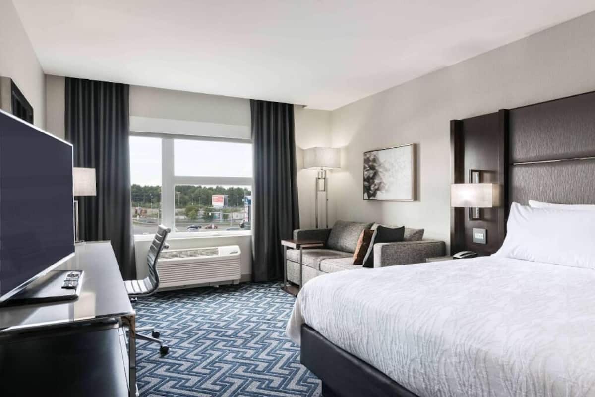 A hotel room with a large bed, a desk with a TV, a sofa, a lamp, and a view of the outside.