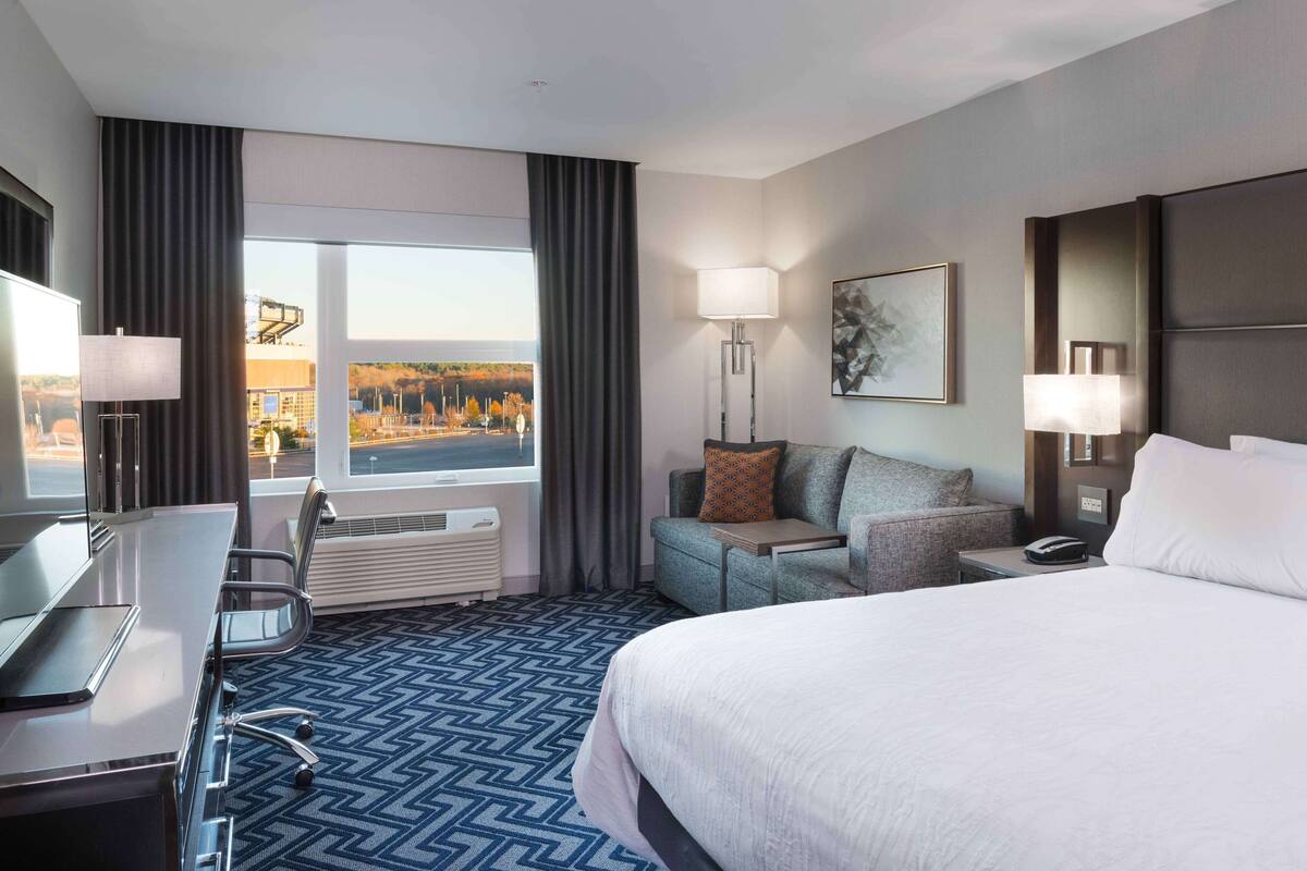Een hotelkamer met een groot bed, een bureau met stoel, een bank, een staande lamp en uitzicht op de stad.
