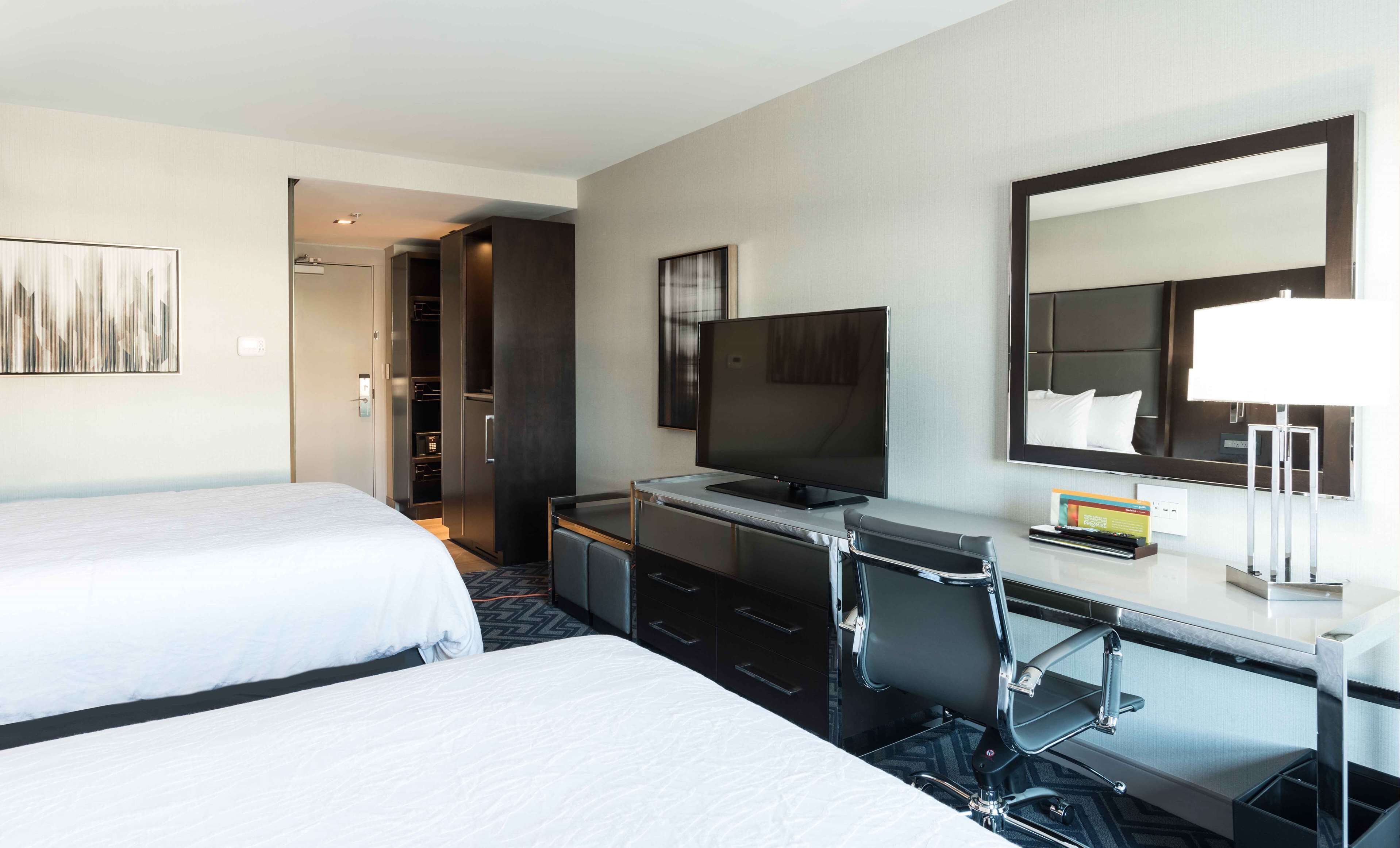 Ein Hotelzimmer mit zwei Betten, einem Schreibtisch mit Stuhl, einem Fernseher, einem Spiegel und einem Kleiderschrank.