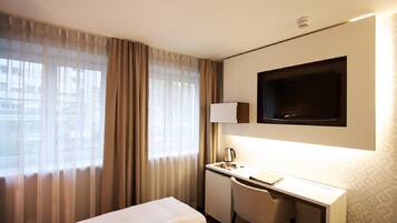 Habitación de hotel con cama, escritorio, silla, televisor y ventana con cortinas.