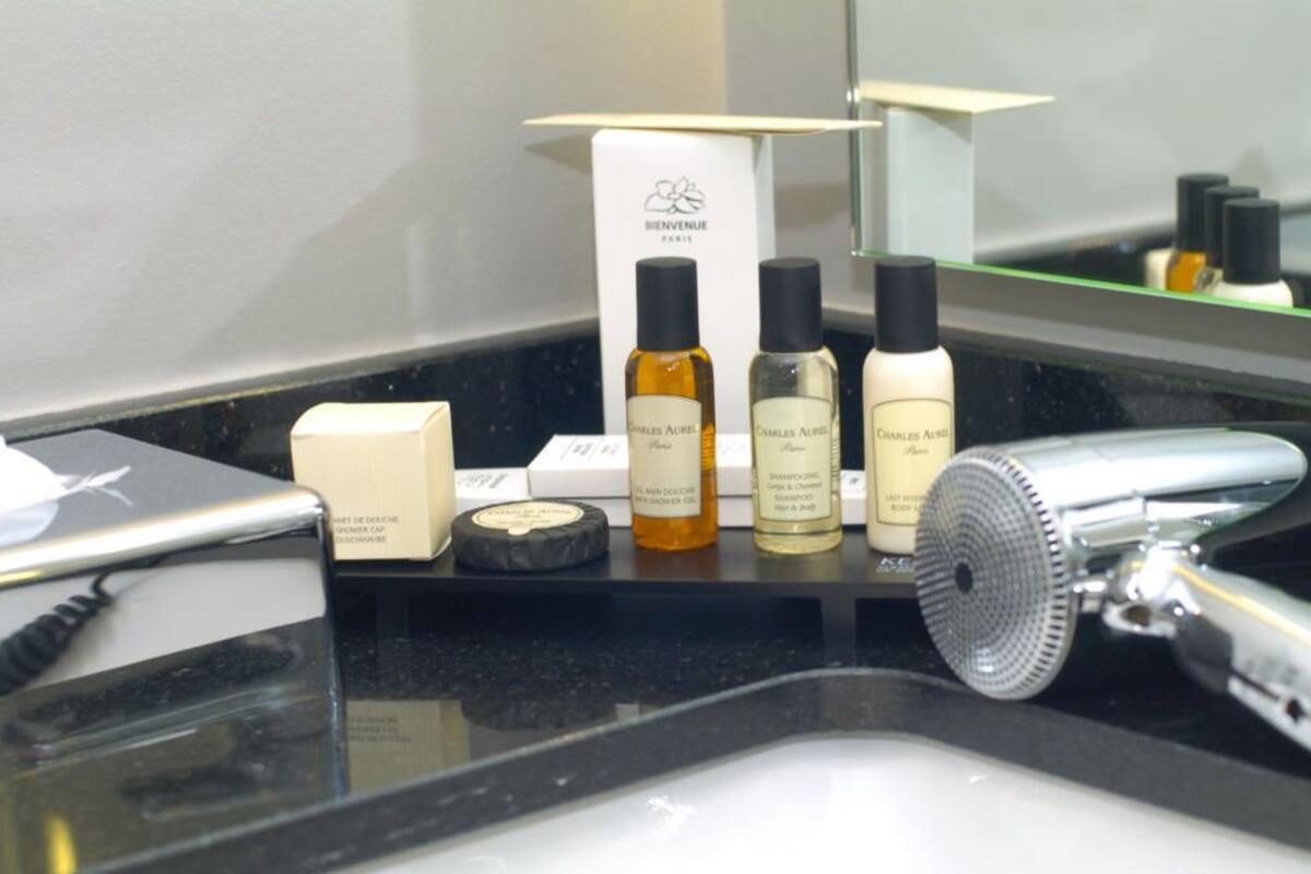 Un meuble-lavabo de salle de bain avec un sèche-cheveux, trois flacons de gel douche Charles Aveline et une boîte de bonnets de douche.