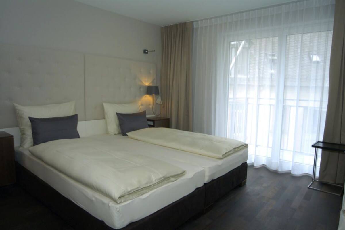 Un lit double avec une tête de lit, une table de chevet, une lampe et une fenêtre avec des rideaux légers.