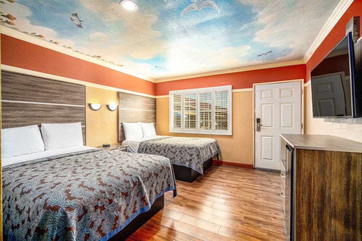 A hotel room with two beds, a wooden floor, a ceiling mural, a TV, and a door.