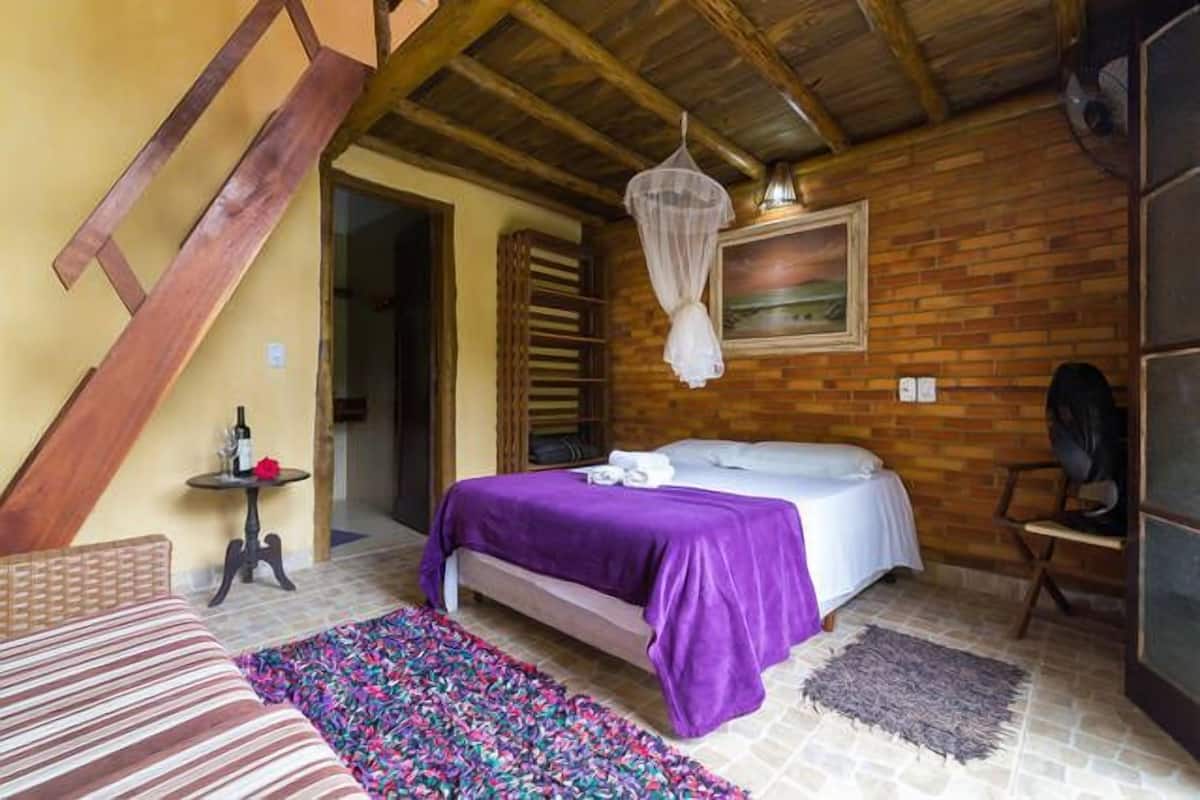 A room with a bed, a chair, a small table with a bottle, and a hanging light fixture.