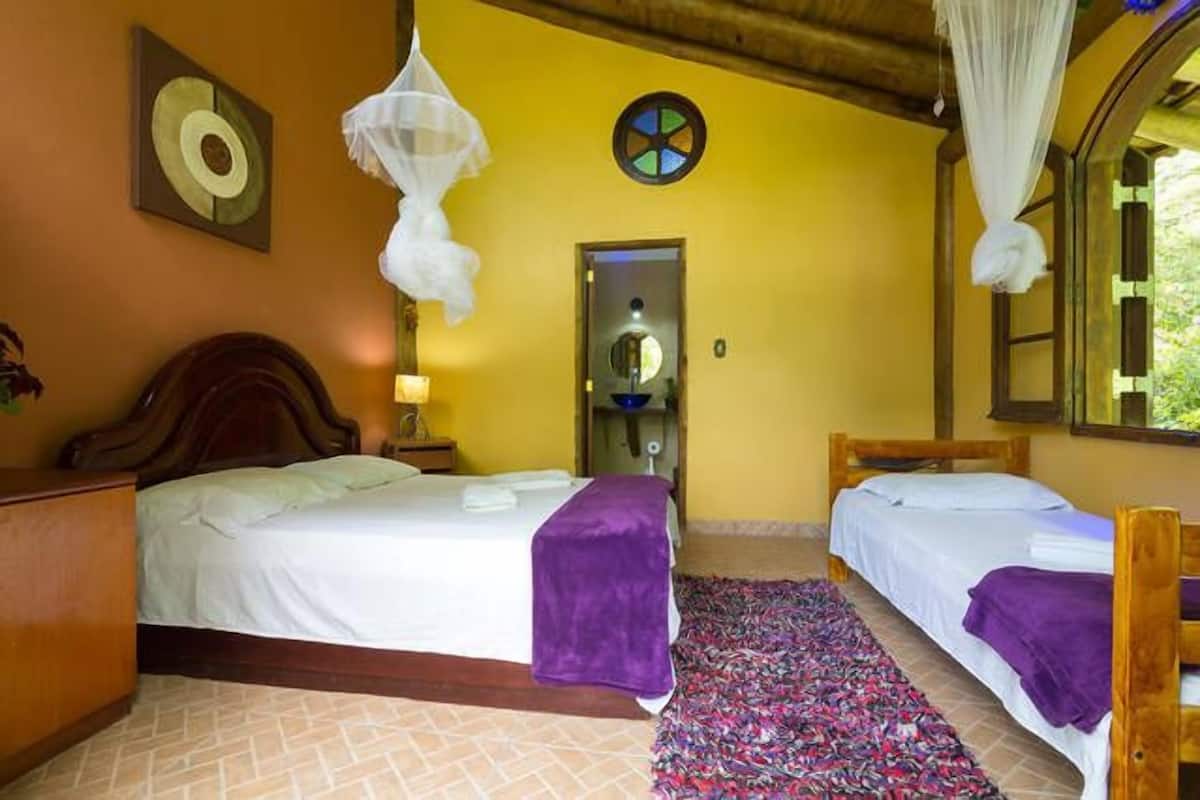 A room with two beds, a wooden headboard, a patterned rug, a wall clock, and a window with a view of greenery.