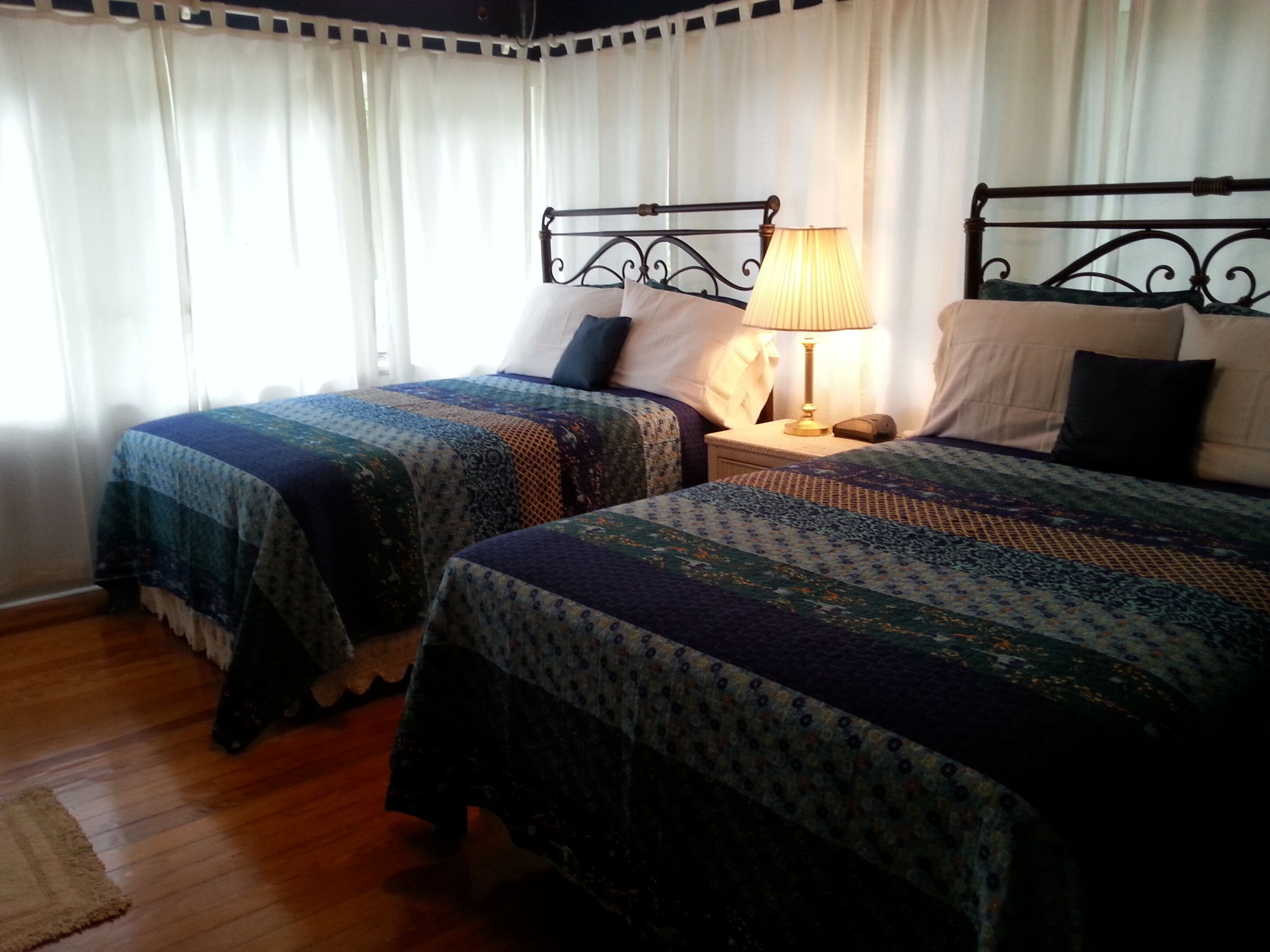 Una habitación con dos camas, cada una con una colcha estampada, una mesita de noche con lámpara y una ventana con cortinas traslúcidas.