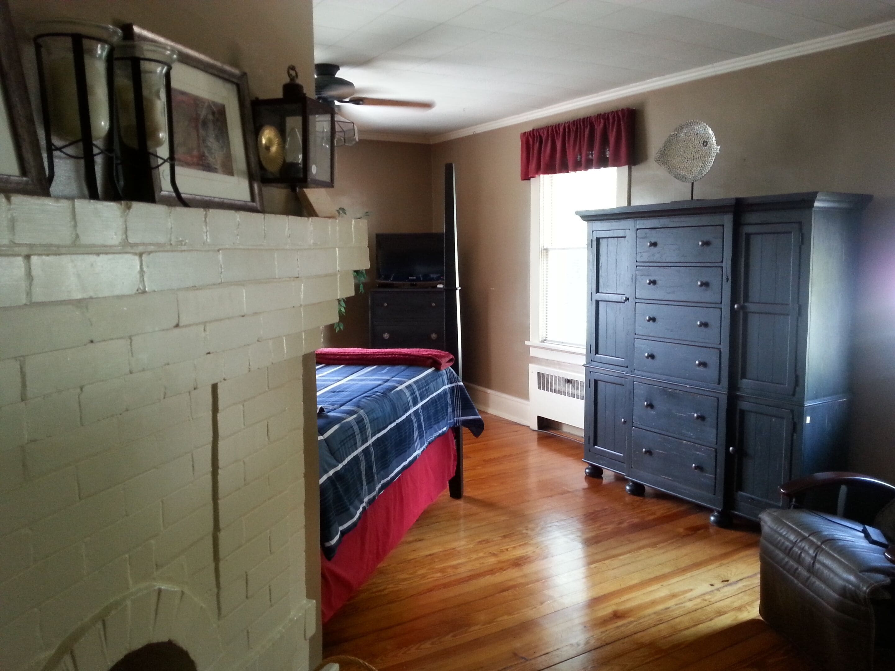 A bedroom with a brick wall, wooden floor, a bed with a red and blue bedspread, a dresser, a television, and a ceiling fan.