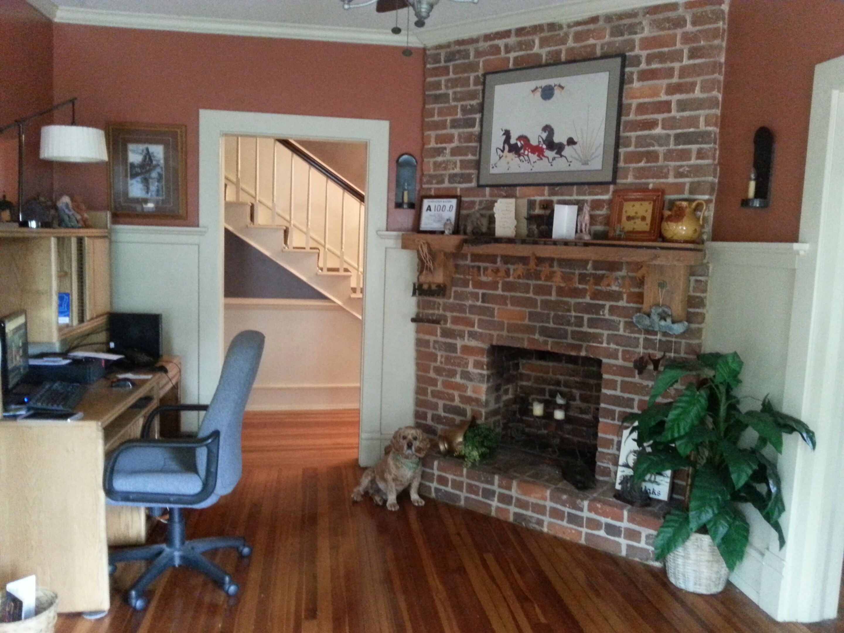 A living room with a brick fireplace, a staircase, a desk with a computer, and a dog.