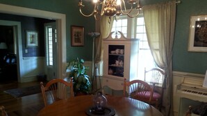 A dining room with a wooden table, chairs, a chandelier, a piano, and a window with curtains. - Hunter House Bed and Breakfast (Madison)