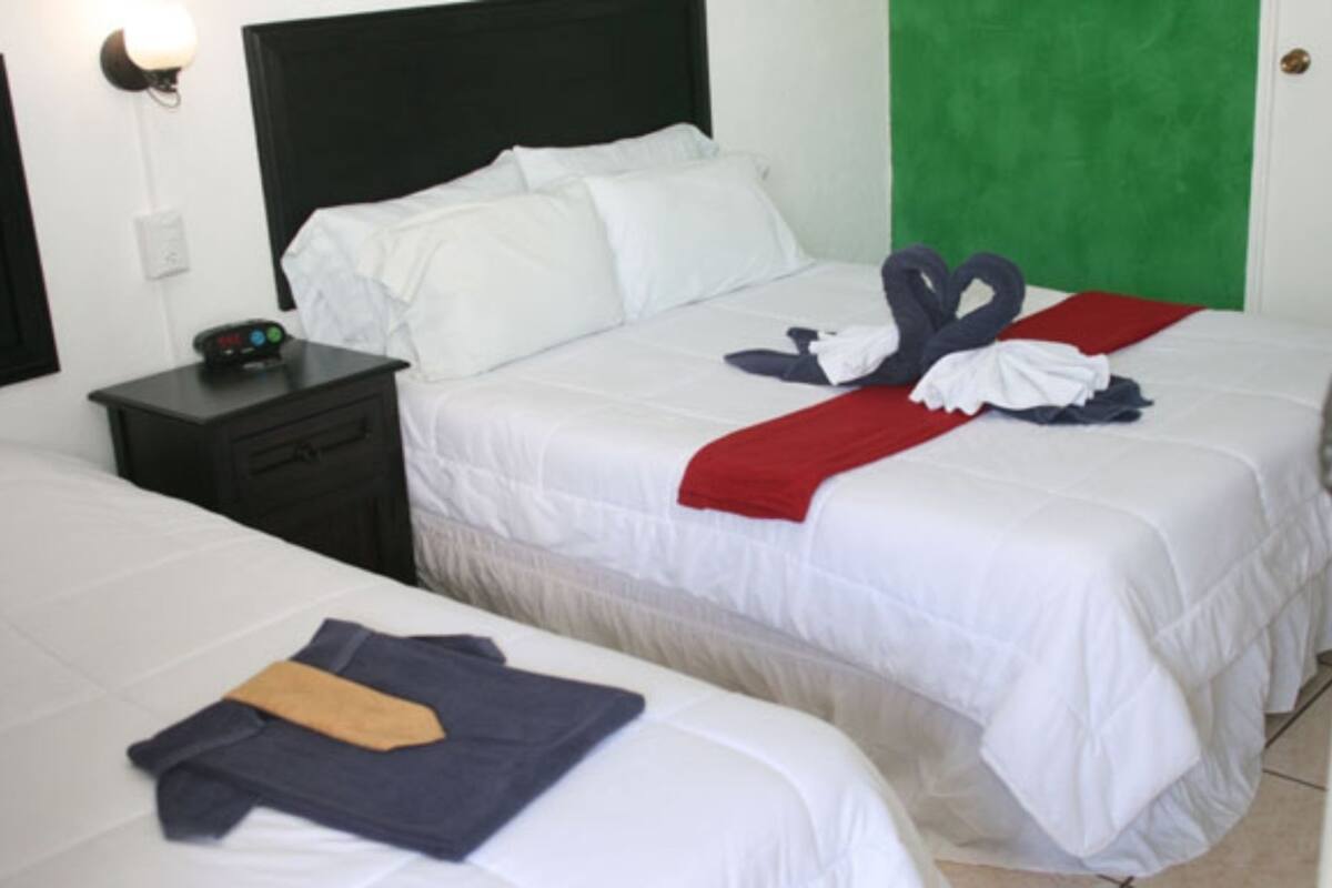 Habitación de hotel con dos camas, cada una con ropa blanca y una manta roja. Hay una mesita de noche con un reloj y un enchufe.