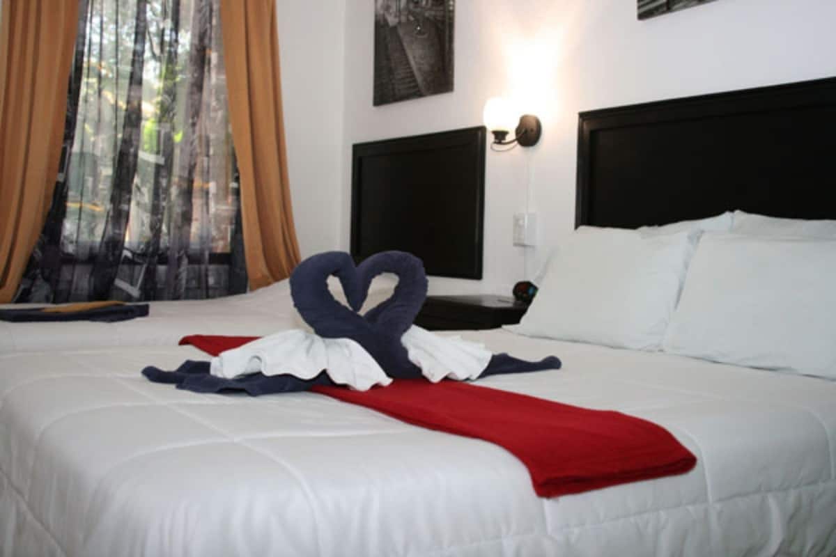 Una cama bien hecha con una toalla dispuesta en forma de corazón, una manta roja y un cabecero negro.