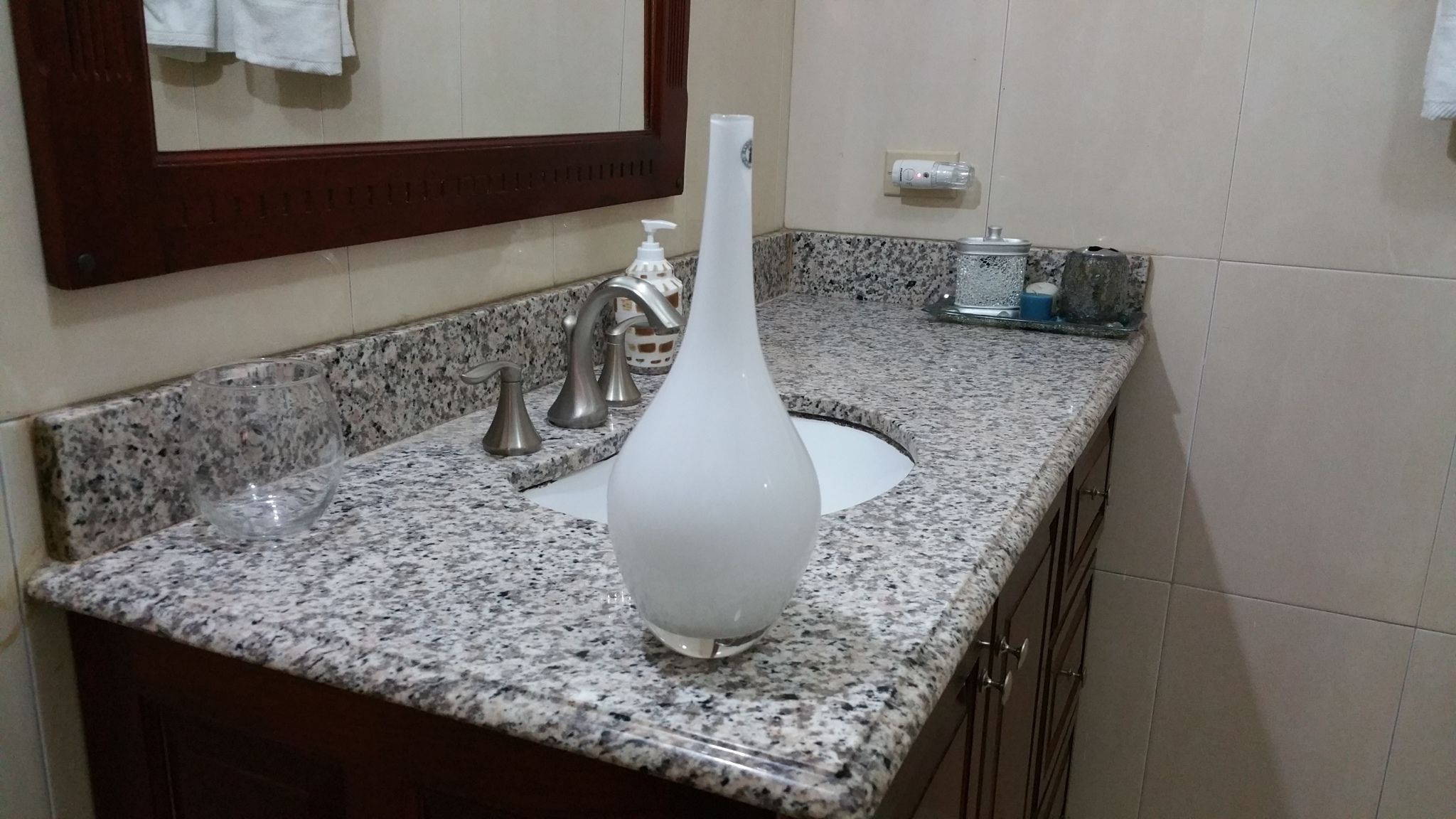 Een badkamer met een granieten aanrechtblad, een witte vaas, een zilveren kraan en een spiegel boven de wastafel.