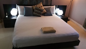 Una cama bien hecha con una toalla doblada, una almohada estampada y dos lámparas de noche en mesitas.