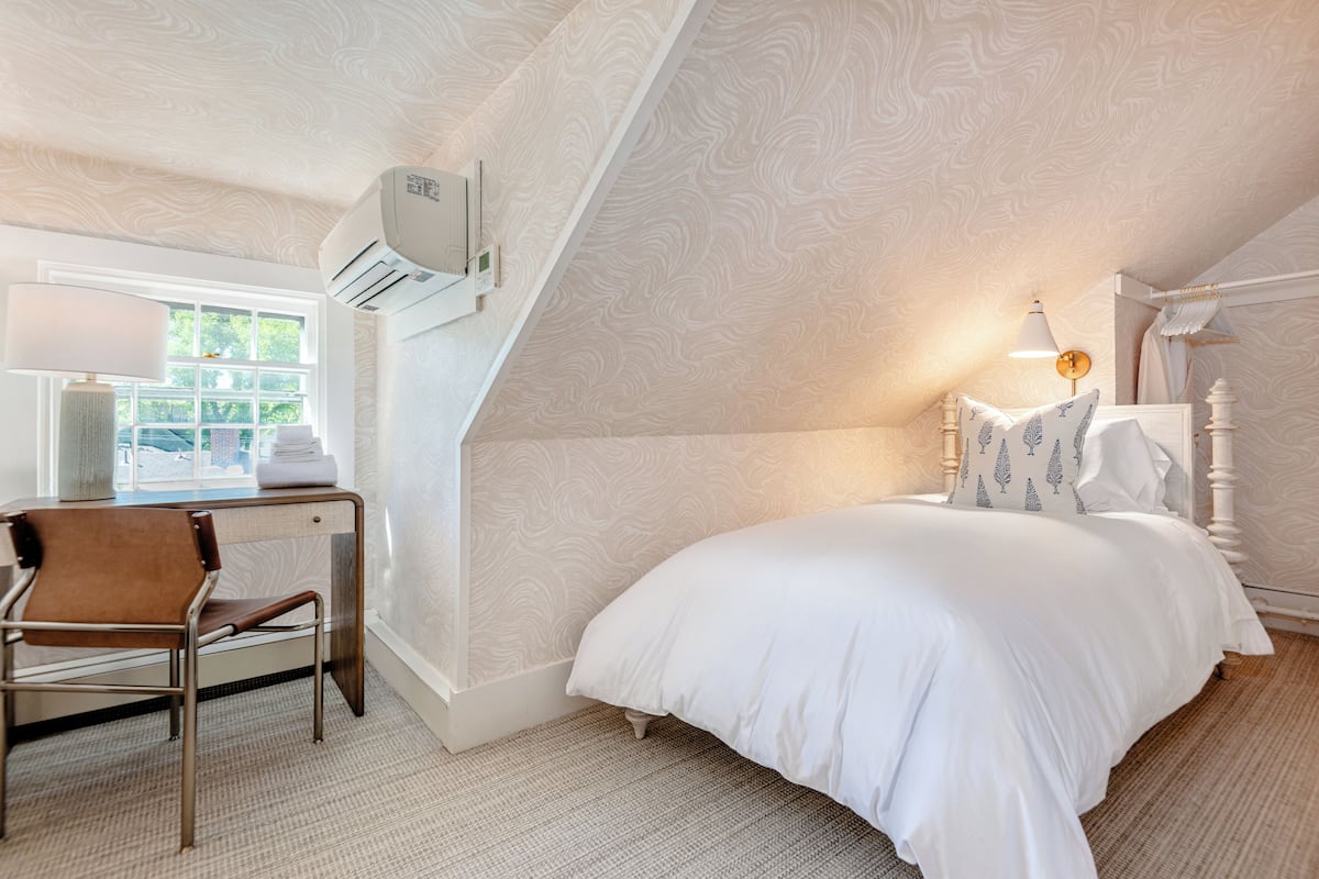 A bedroom with a bed, a desk with a chair, a window, and an air conditioning unit.