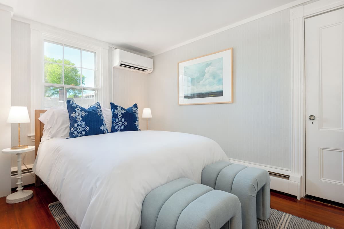 A spacious bedroom with a large bed, a bedside table with a lamp, a window with blinds, and a door.