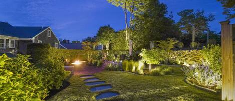 A well-lit garden pathway with stepping stones, surrounded by lush greenery and a stone wall.