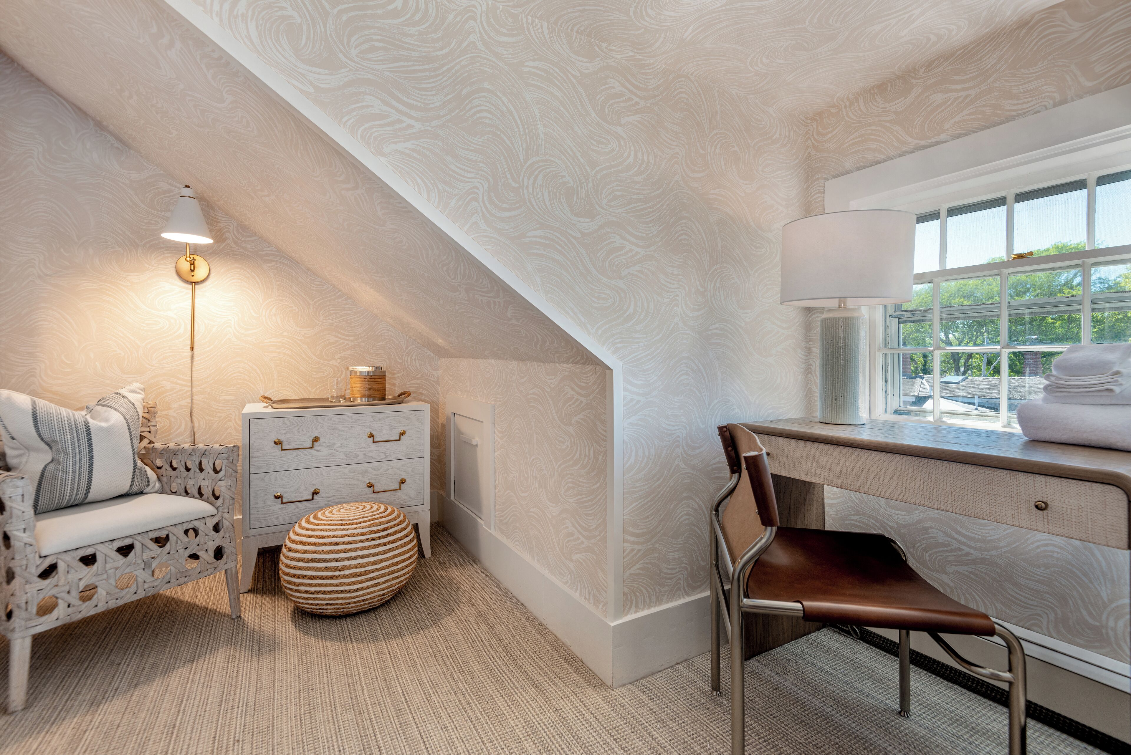 A cozy room with a white patterned ceiling, a white dresser with drawers, a wicker ottoman, a brown leather chair, and a window with a view of greenery.