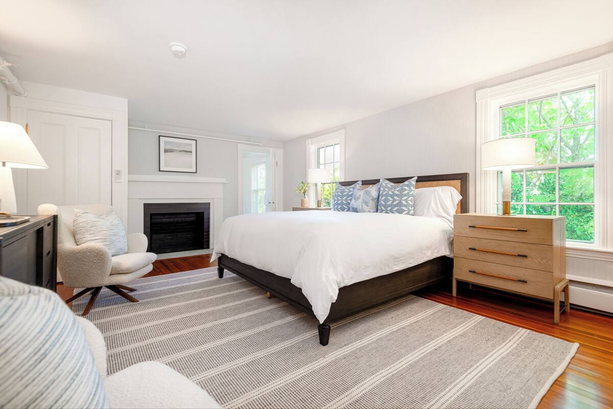 A bedroom with a large bed, a dresser, a chair, a fireplace, and a window with a view of trees.
