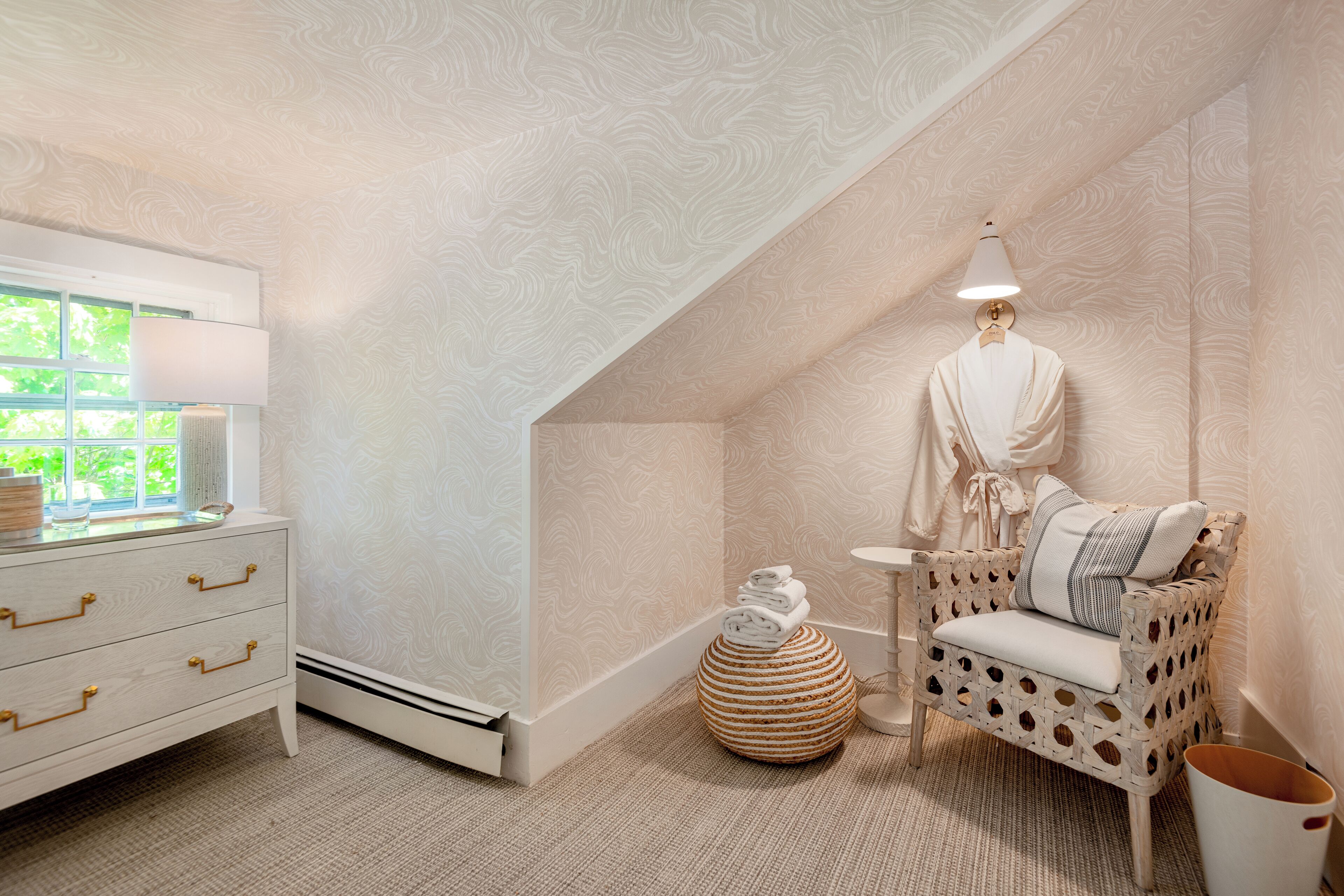 A cozy room with a white dresser, a wicker chair with cushions, a small round table, and a lamp.