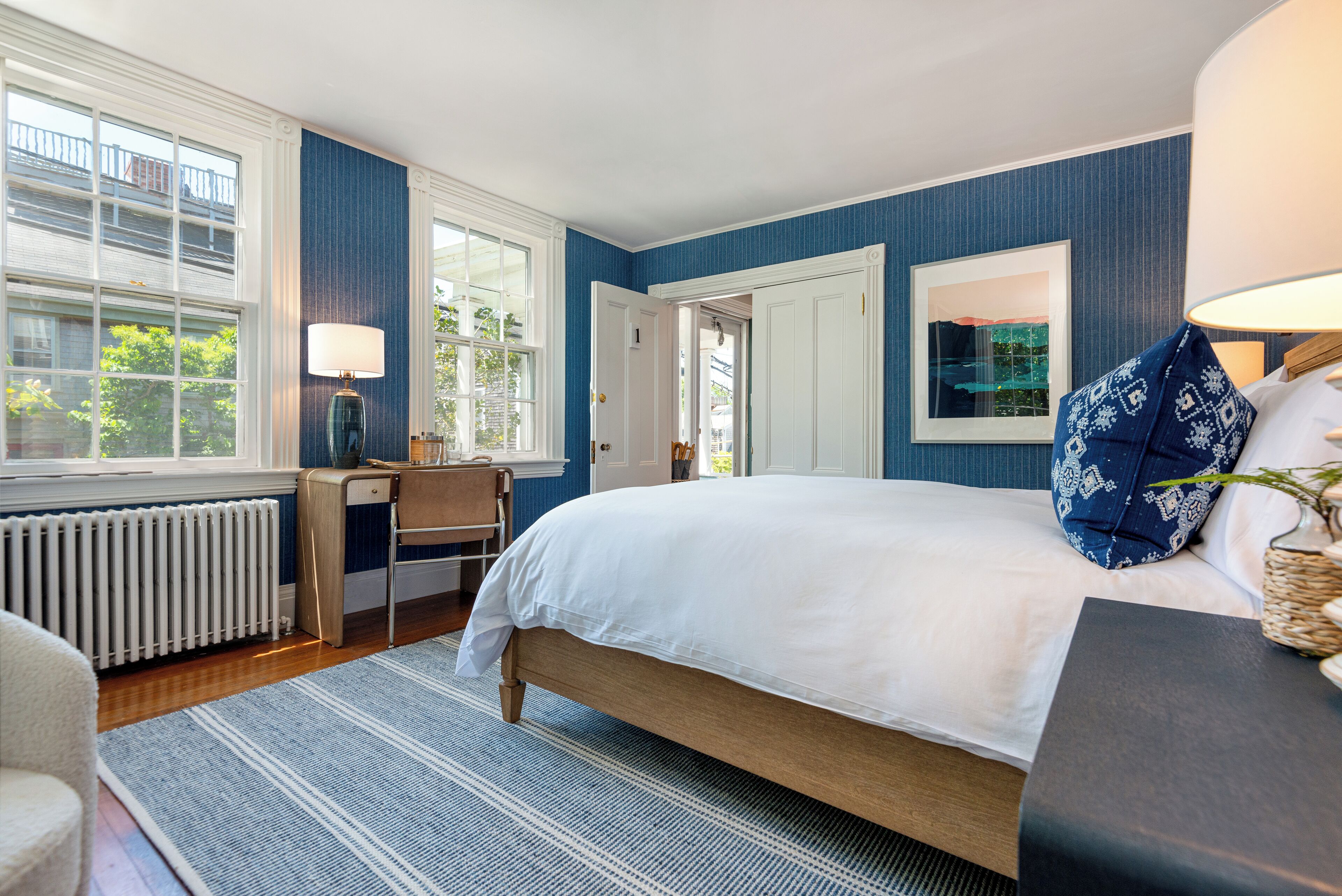 A bedroom with a large bed, a desk, and a chair. There is a window with a view of trees and a door leading to another room.