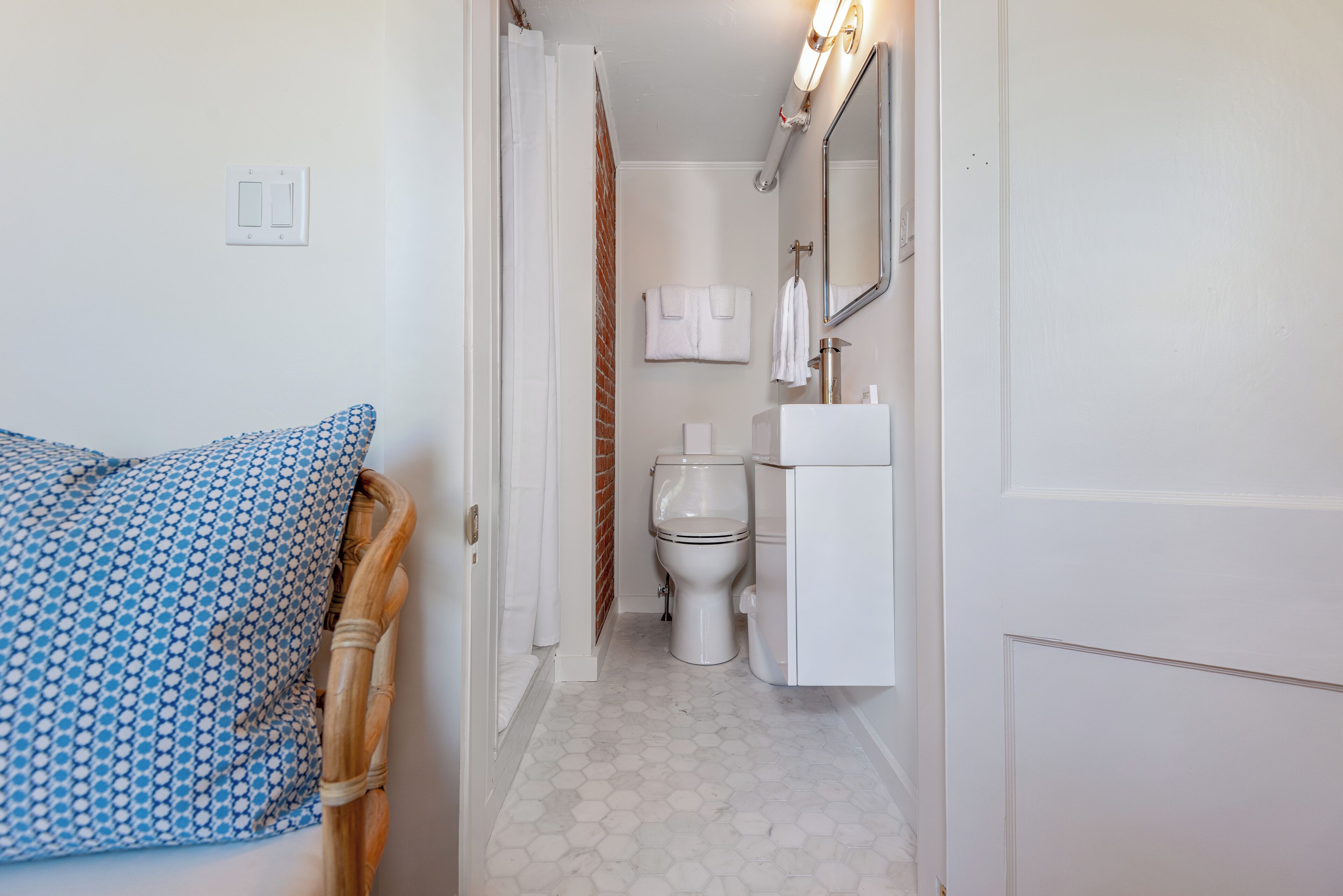 A narrow hallway leading to a bathroom with a toilet, sink, and a mirror. There is a patterned pillow on a chair to the left.