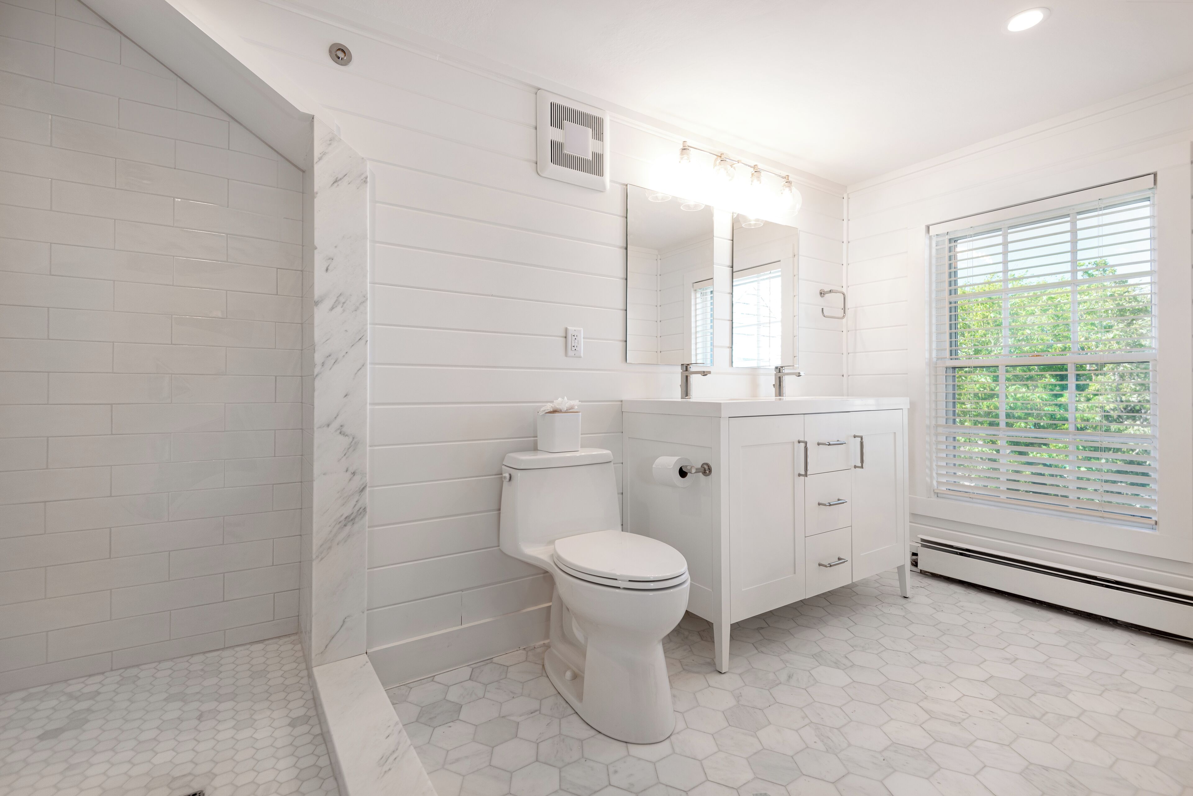 A modern bathroom with white tiles, a toilet, a vanity, and a large window.