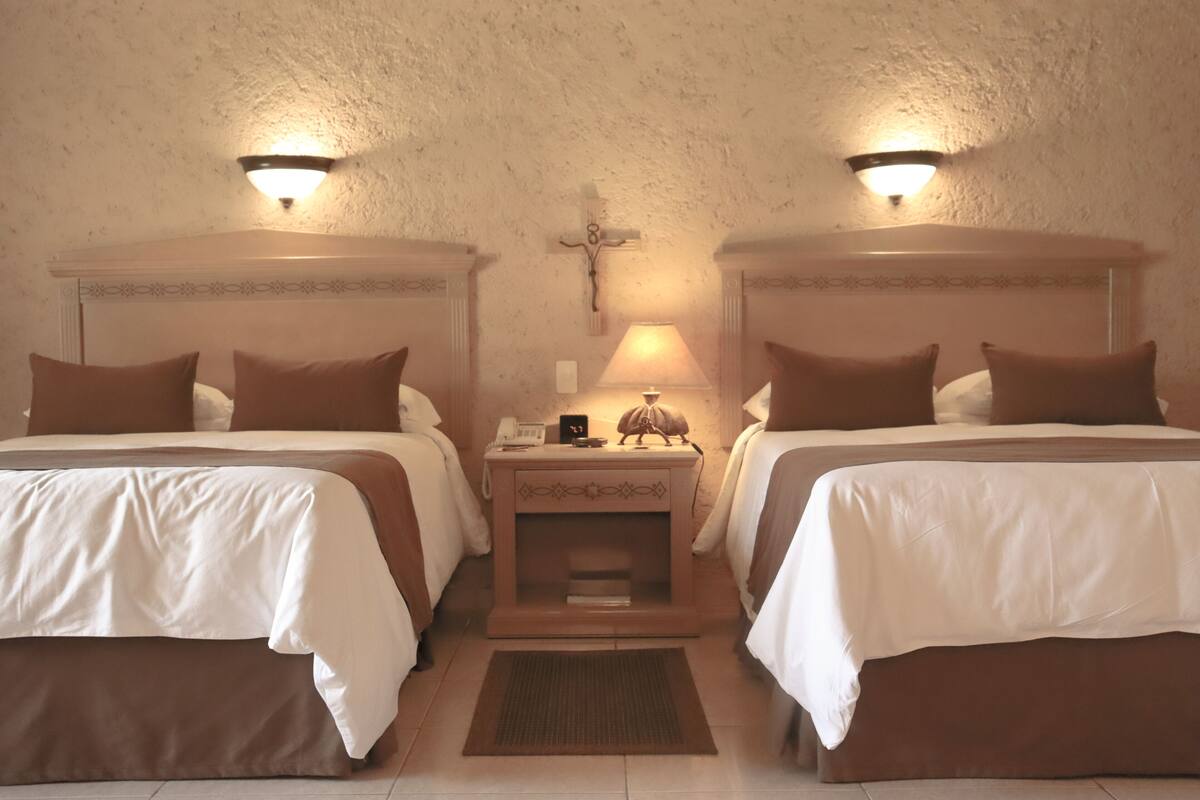 Une chambre d’hôtel avec deux lits, chacun doté d’une tête de lit, d’une table de chevet avec une lampe et d’une croix accrochée au mur.