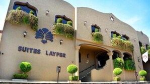 Een gebouw met het bord 'SUITES LAYFER', wat een hotel aangeeft. Er zijn meerdere ramen met beplanting, een trap en een heldere lucht.