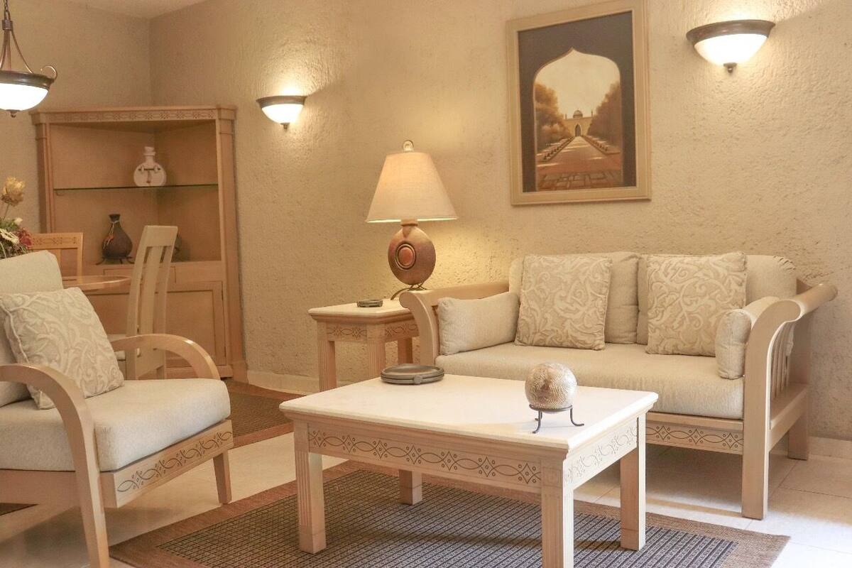 Un salon avec un canapé beige, deux fauteuils, une table basse, une lampe, une étagère ornée d’objets décoratifs et un tableau encadré au mur.
