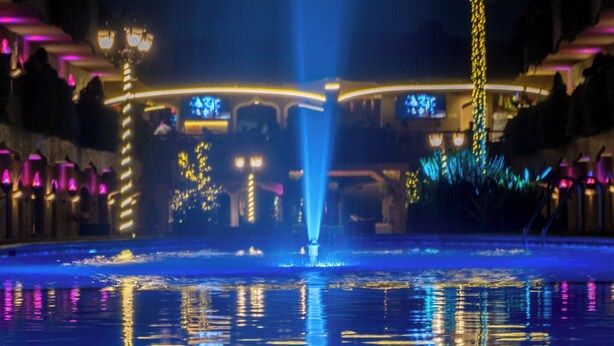 ’s Nachtzicht op een fontein met waterstralen en reflecties op een nat oppervlak, omringd door verlichte gebouwen en palmbomen.