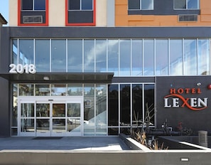 Uma entrada de hotel moderna com fachada de vidro, um ano exibido acima da porta e o nome 'HOTEL LEXEN' à direita.