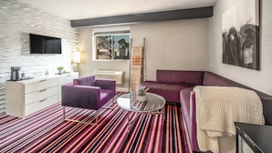 A modern living room with a striped carpet, a purple sofa, a glass coffee table, a white dresser, and a flat-screen TV mounted on a textured wall. - Lexen Hotel- Hollywood Walk of Fame/Universal Studios (Los Angeles)