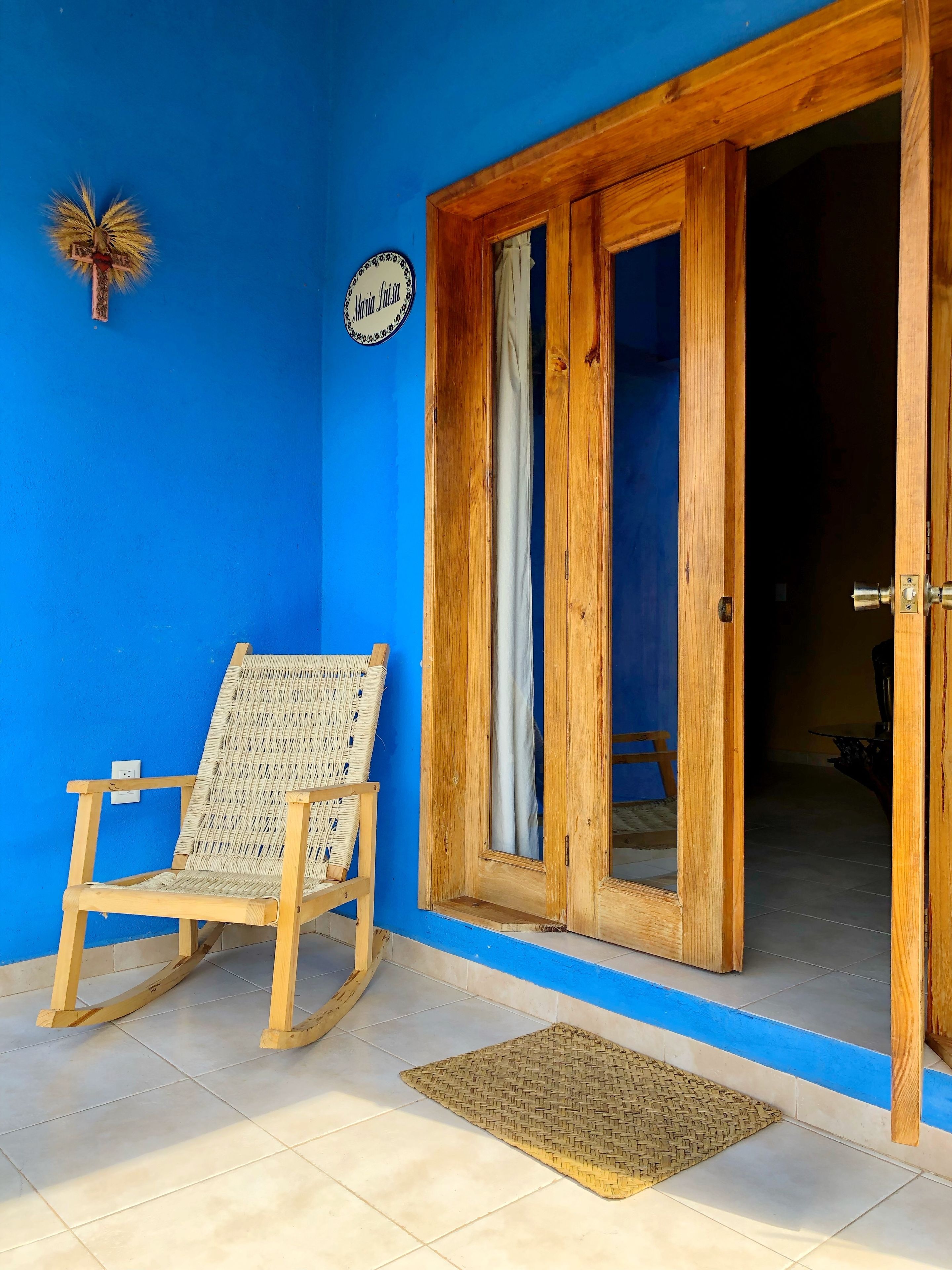 Una puerta de madera con un panel de vidrio, una mecedora de mimbre y un felpudo sobre suelo de baldosas.