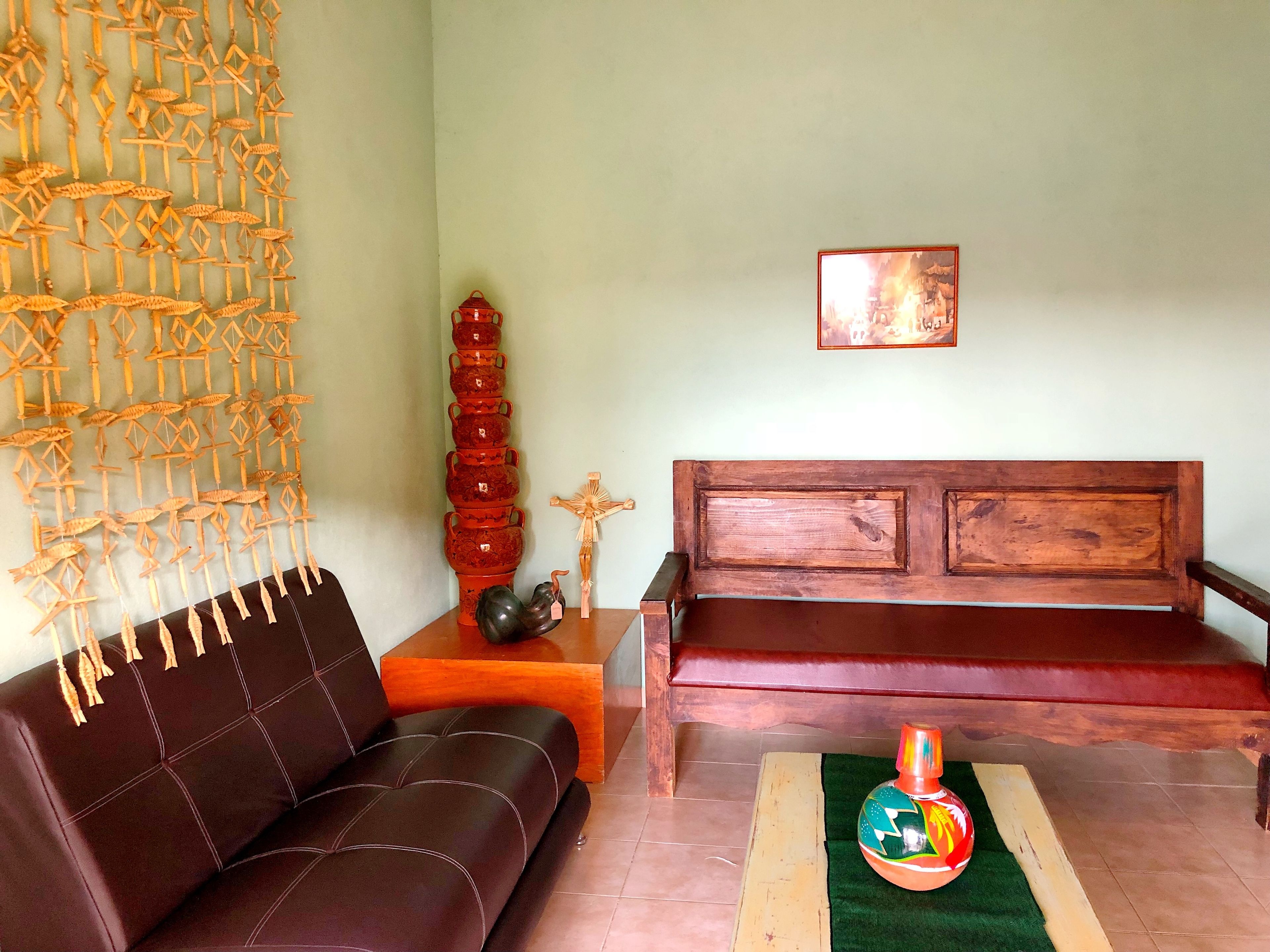 Ein Wohnzimmer mit einer braunen Ledersofa, einer Holzbank, einem Wandbehang, einem hohen Stapel roter Vasen, einem hölzernen Kruzifix und einem gerahmten Bild an der Wand.