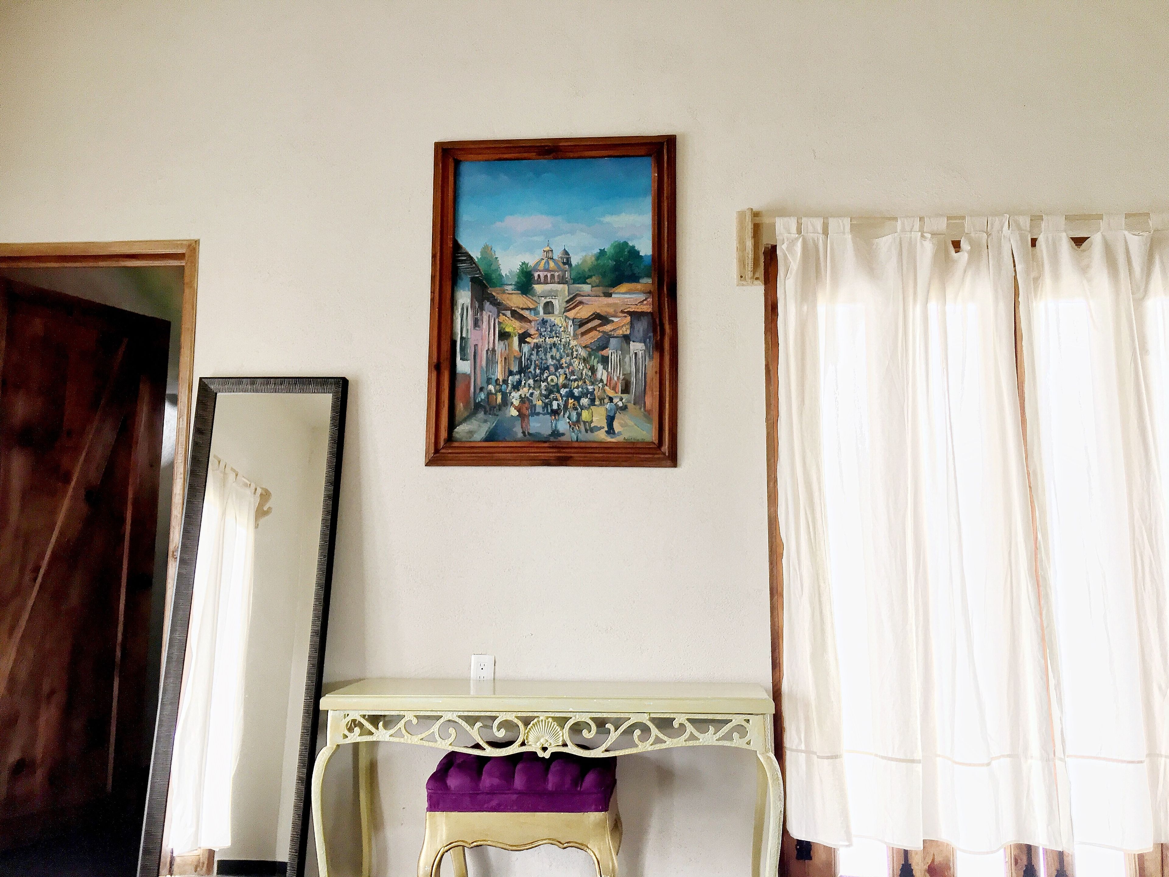 Una habitación con un cuadro de una escena callejera, un espejo, una mesa con un cojín morado y cortinas blancas.