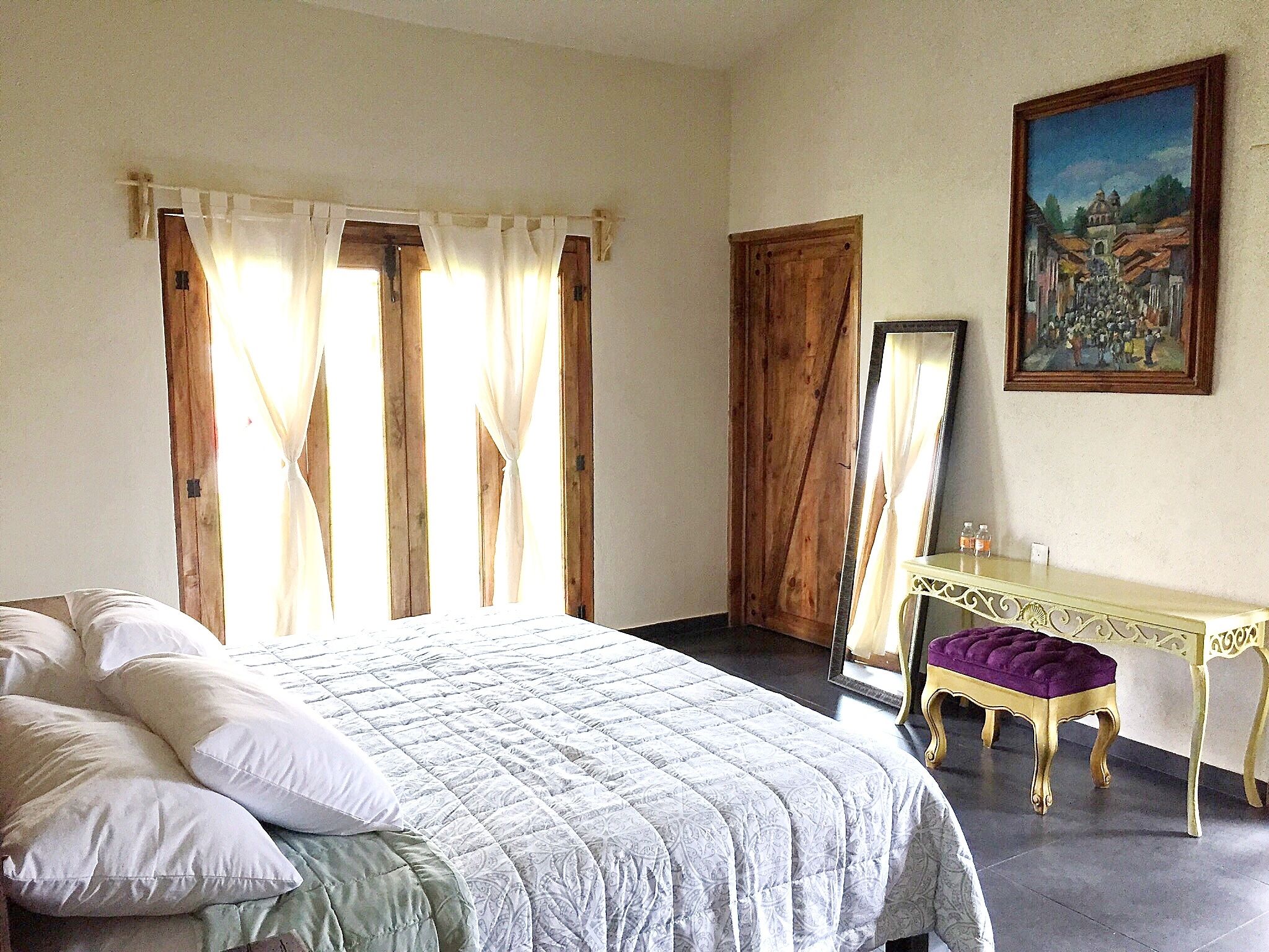 Een slaapkamer met een bed, gordijnen, een houten deur, een spiegel, een klein tafeltje en een schilderij aan de muur.