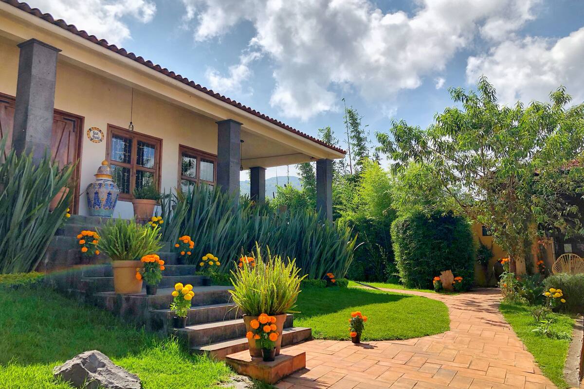 Una casa con porche cubierto, escalones que llevan a la entrada y un jardín con plantas y flores en macetas.