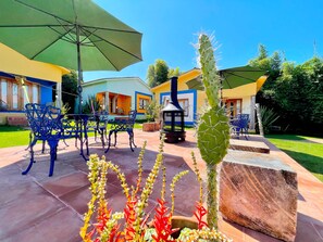 Un patio al aire libre con sillas de metal azules, sombrillas verdes, un cactus y un edificio con paredes azules y amarillas.
