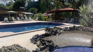 A swimming pool with a tiled deck, surrounded by rocks and a waterfall feature. There are lounge chairs and umbrellas in the background. - Hotel Manuel Antonio Park (Quepos)
