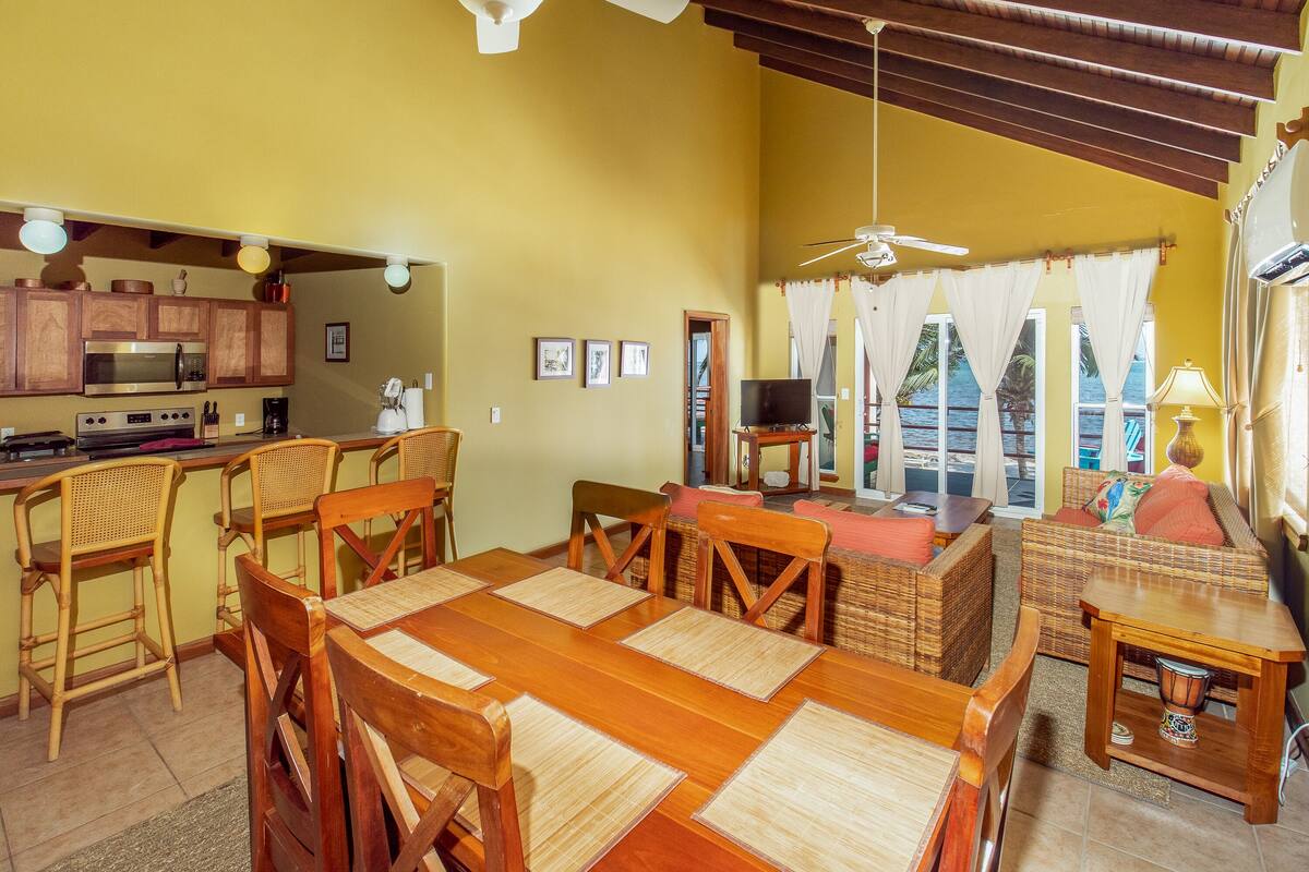 A spacious living area with a dining table, chairs, a sofa, and a television. There is a kitchen area with a microwave and a coffee maker. The room has a ceiling fan and large windows with curtains.