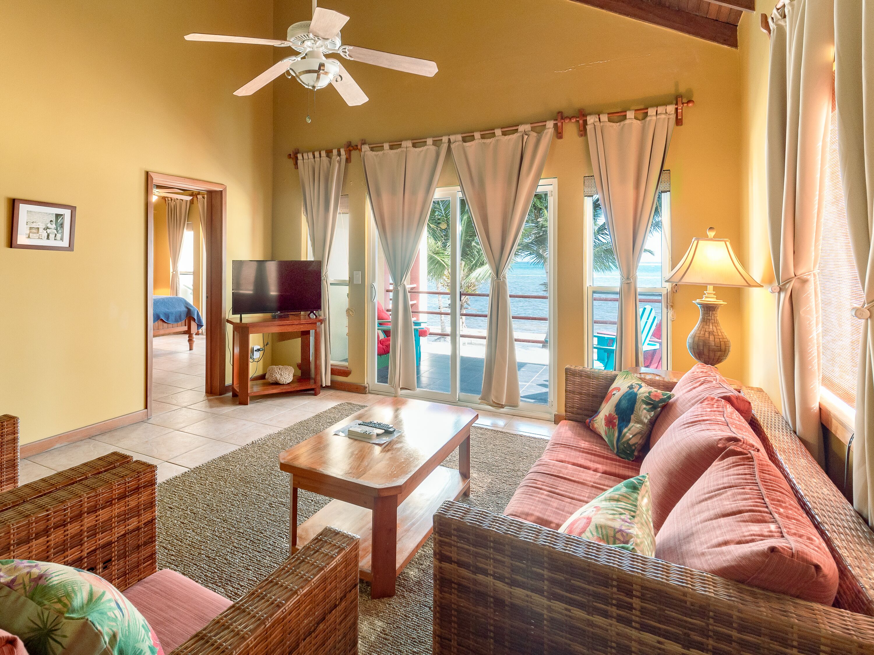 A living room with wicker furniture, a television, and a view of the sea through large windows.