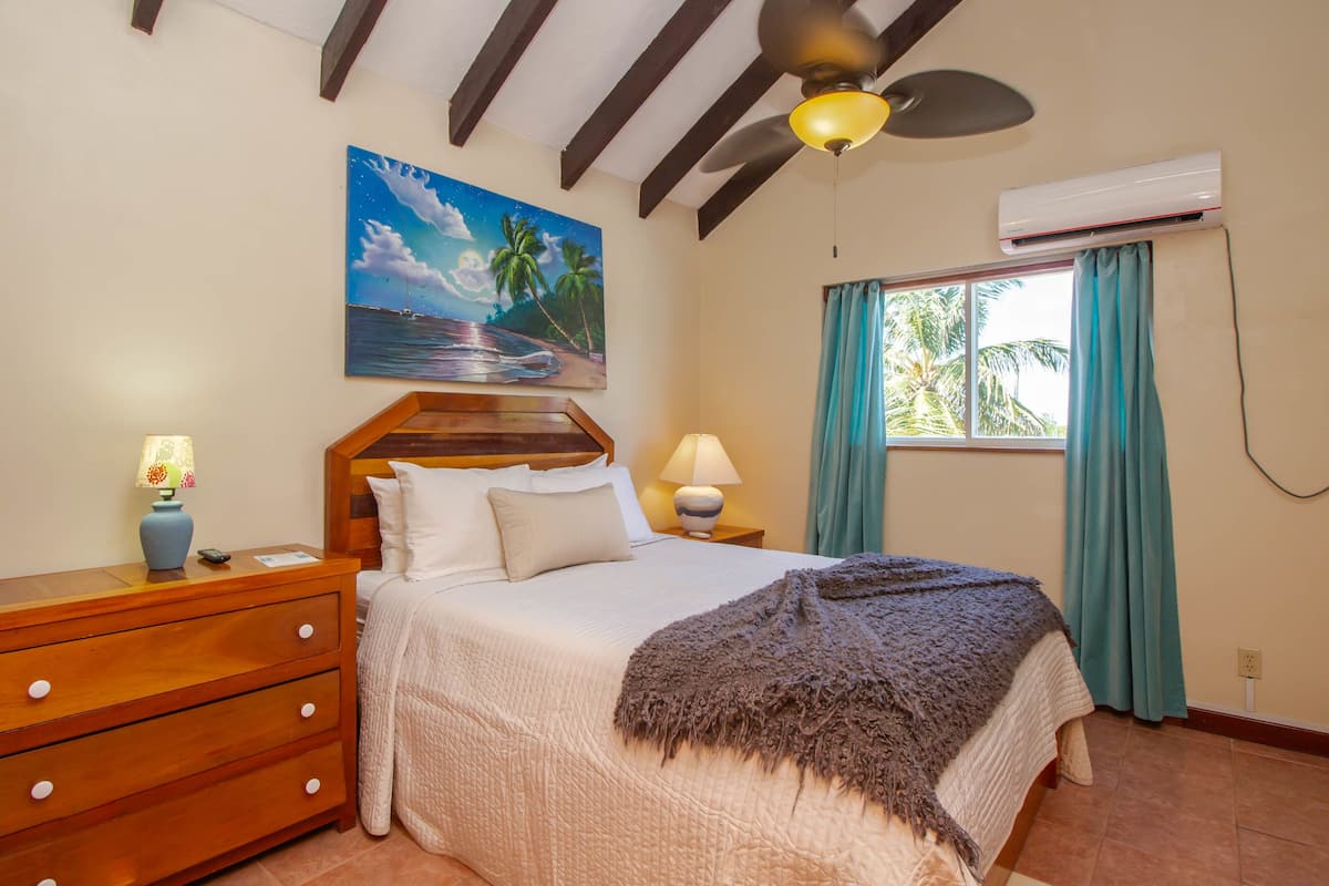 A bedroom with a bed, wooden nightstands, a ceiling fan, a painting, and an air conditioning unit.