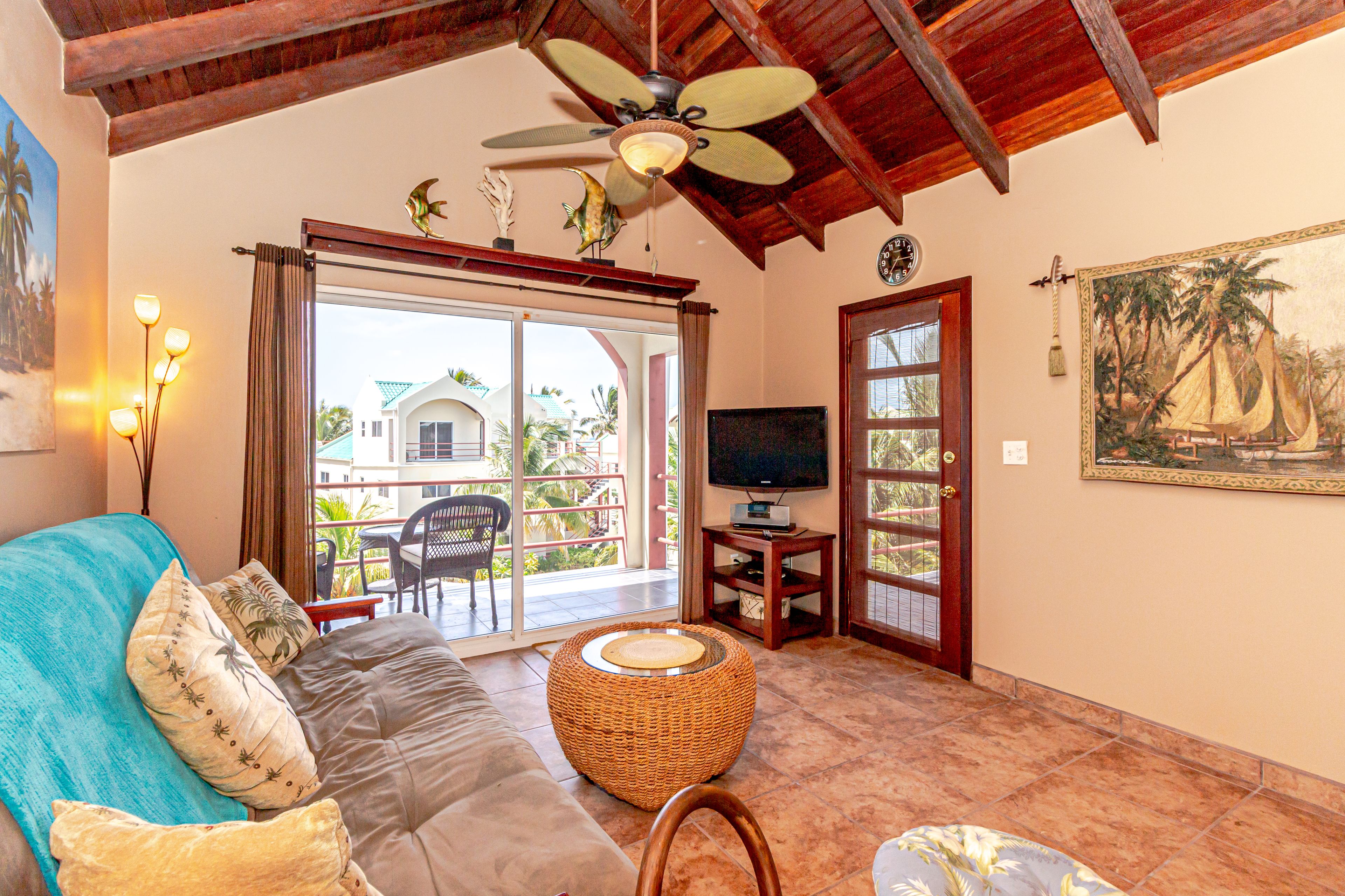 A living room with a sofa, a television, and a balcony with a view of palm trees.