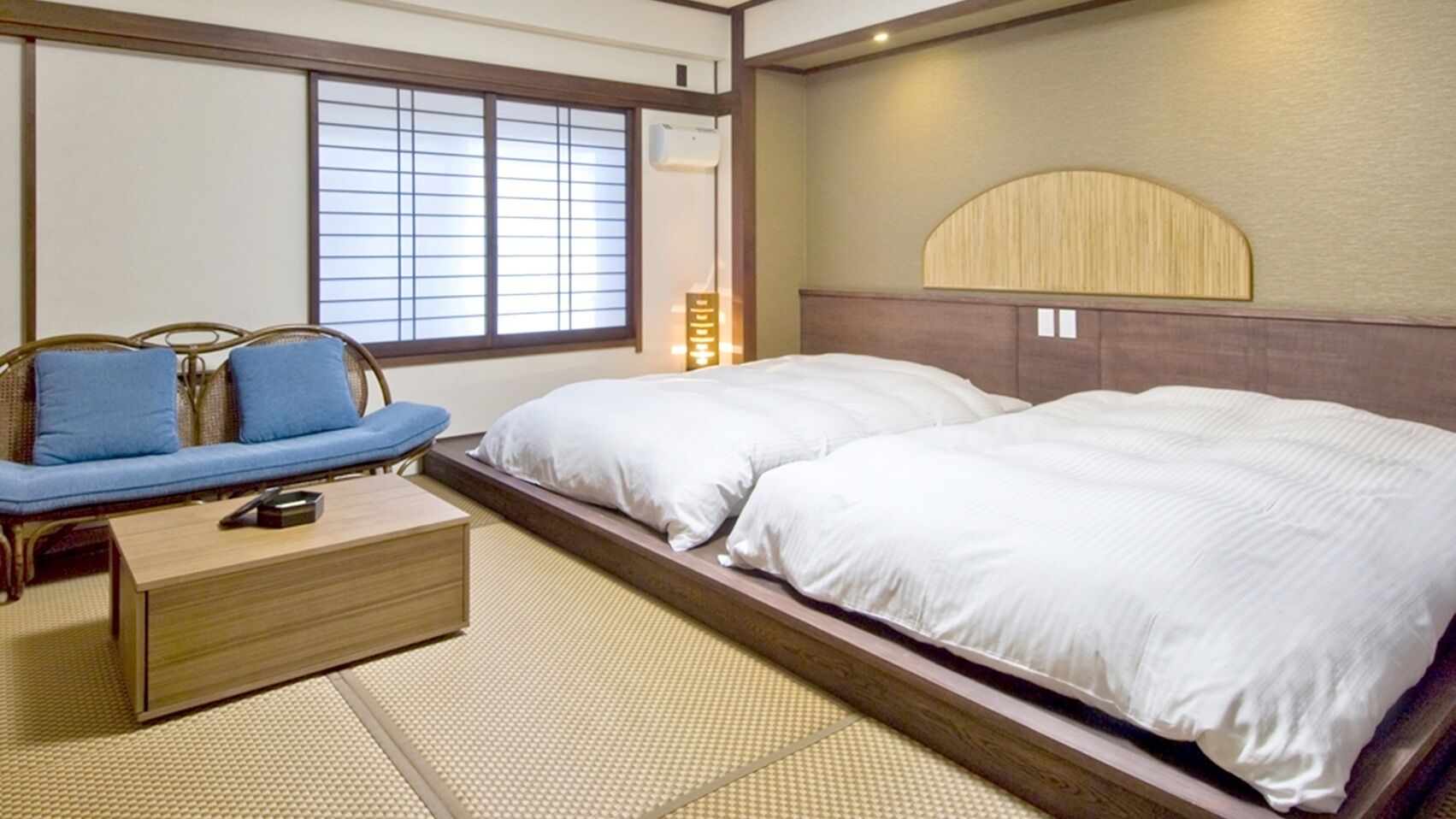 Una habitación de estilo japonés tradicional con suelo de tatami, una cama baja de madera con ropa blanca, un sofá azul con cojines y una pequeña mesa de madera.