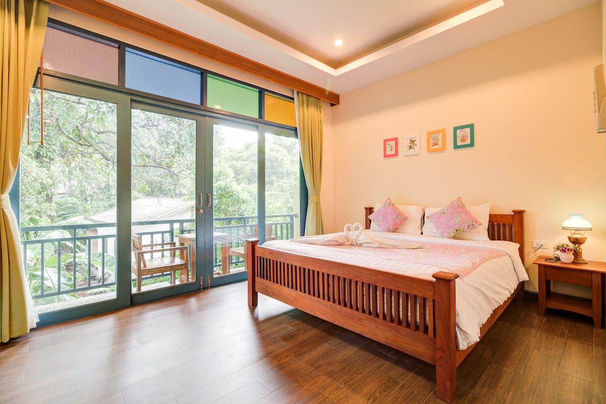 A bedroom with a wooden bed, a balcony with a chair, and a view of greenery.