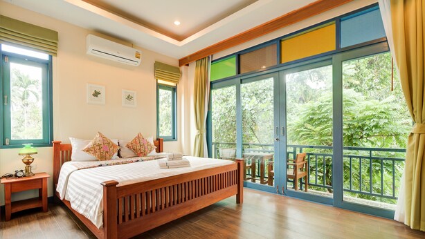 A bedroom with a wooden bed, a nightstand, a window with curtains, and a view of greenery outside. - Baan Patcharintorn Samui (Koh Samui)